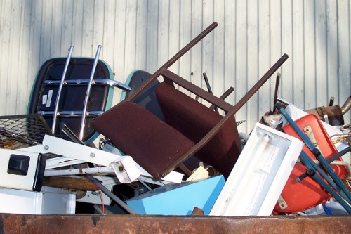 A pile of discarded household and garden items located outdoors against a vertical metal sheet fence. The heap includes a variety of materials such as a dark brown fabric-upholstered chair with wooden legs, a light blue plastic container, a white window frame, a black metal mesh basket, and an orange plastic toolbox. Some items are stacked or leaning on each other, with the chair's legs sticking upward. The background features a corrugated metal fence with a weathered appearance, and the scene is lit with natural daylight, highlighting the different textures and colors of the debris. This collection of rubbish likely represents waste that would be collected for removal services, similar to those offered by Garden Clearance Downham, who specialise in rubbish clearance in the area near Downham in the UK.
