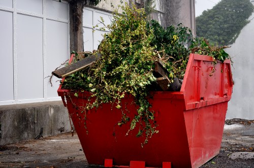 A large red plastic skip filled with garden debris including leafy green branches, stems, and shrub cuttings, positioned on a paved outdoor area beside a white fence and a concrete wall, with some garden tools and small wooden planks visible among the waste, in a natural daylight setting that suggests a garden clearance or rubbish removal cleanup, typical of services provided by Garden Clearance Downham in the local area of Downham, showing an organized collection of garden waste ready for disposal or collection.
