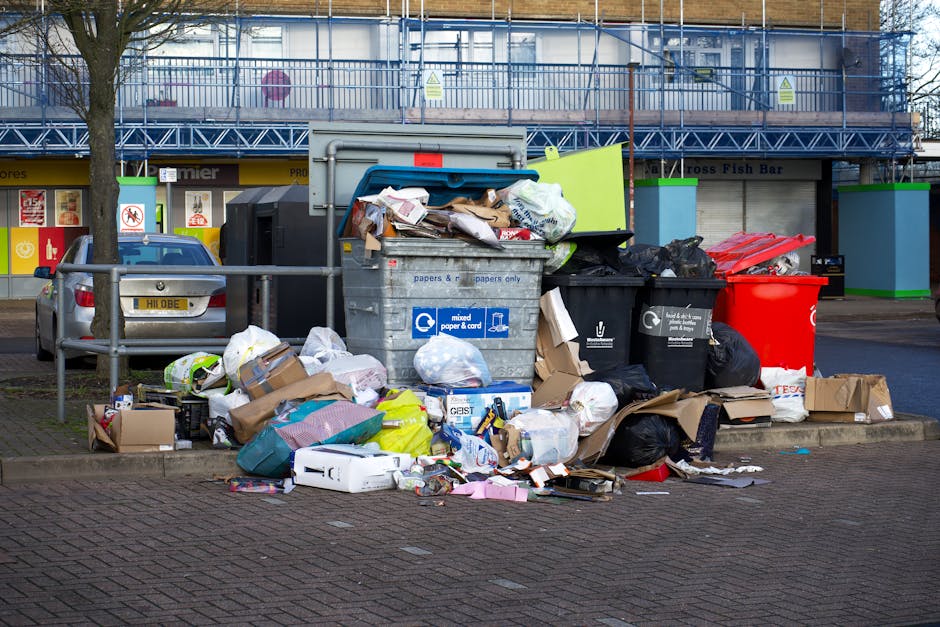 A black wheeled bin labeled 'St. John's' positioned on the edge of a residential street pavement at night, filled with various types of bulky rubbish including flattened cardboard boxes, plastic packa