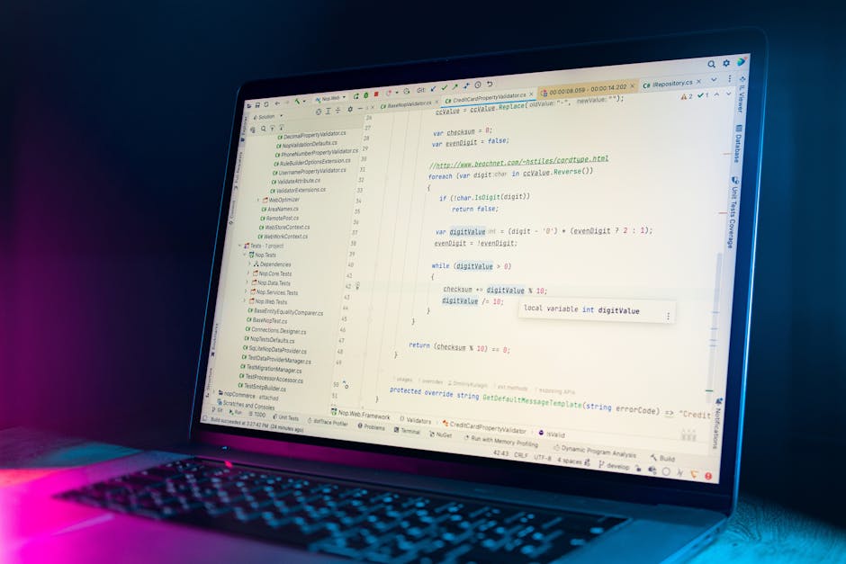 A close-up view of a person's hands operating a laptop with a dark-themed code editor open on the screen, displaying lines of multicolored programming code. The laptop is positioned on a black table, 