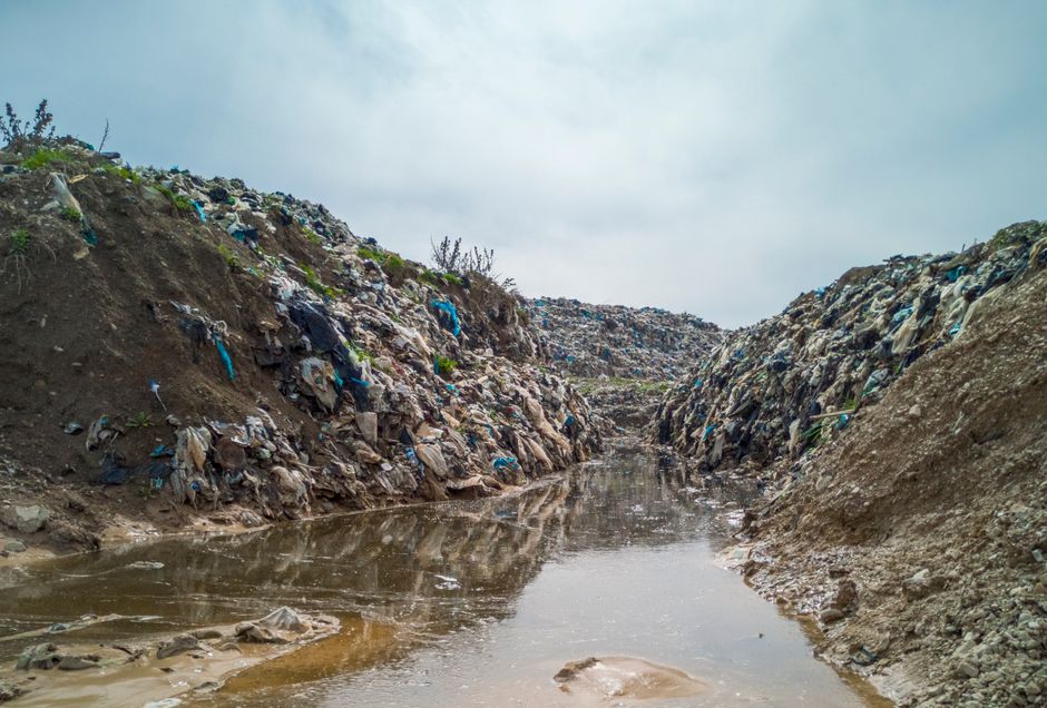 A large collection of waste and rubbish is piled around a communal recycling station on a paved urban street. The pile includes various black, green, and red disposal bins, some overflowing with trash
