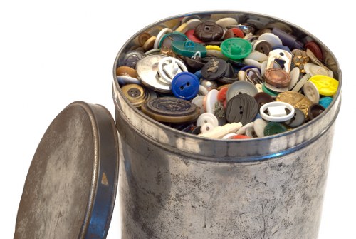 The image shows a round, metal rubbish bin filled with a variety of plastic and metal buttons of different sizes, shapes, and colours, including blue, black, white, red, yellow, and brown. The buttons have various finishes, with some appearing shiny and smooth, while others are matte or textured. The bin is positioned outdoors on a concrete or paved surface, with the lid placed to the side, leaning slightly against the bin's side. The scene appears well-lit, likely by natural daylight, and the buttons are piled haphazardly inside the bin, indicating a collection of discarded small household items. This image relates to rubbish removal services provided by Garden Clearance Downham, particularly in the context of waste disposal or recycling of unwanted household materials, including plastic and metal objects, and is representative of typical rubbish collection tasks in the Downham area.